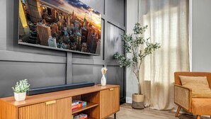 Un soggiorno moderno con una TV a schermo piatto montata a parete, un mobile in legno con libri e una pianta, una sedia in vimini e un'ampia finestra con tende leggere.