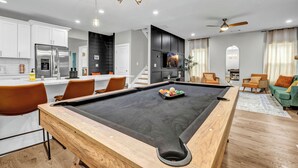 Un soggiorno moderno con tavolo da biliardo, cucina con mobili bianchi e zona pranzo con sedie arancioni.