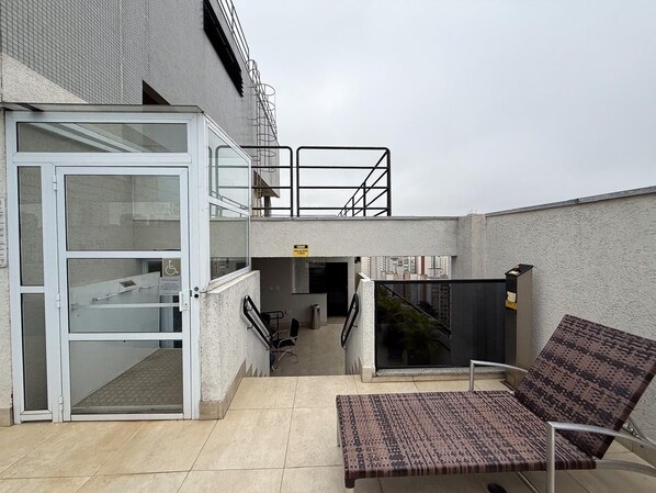 A rooftop terrace with a glass door entrance, a small seating area with a patterned cushion, and a staircase leading to an upper level.