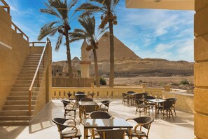 A terrace with tables and chairs, a staircase, and a view of a pyramid. - Secrets Of The Pyramids Hotel (Giza)