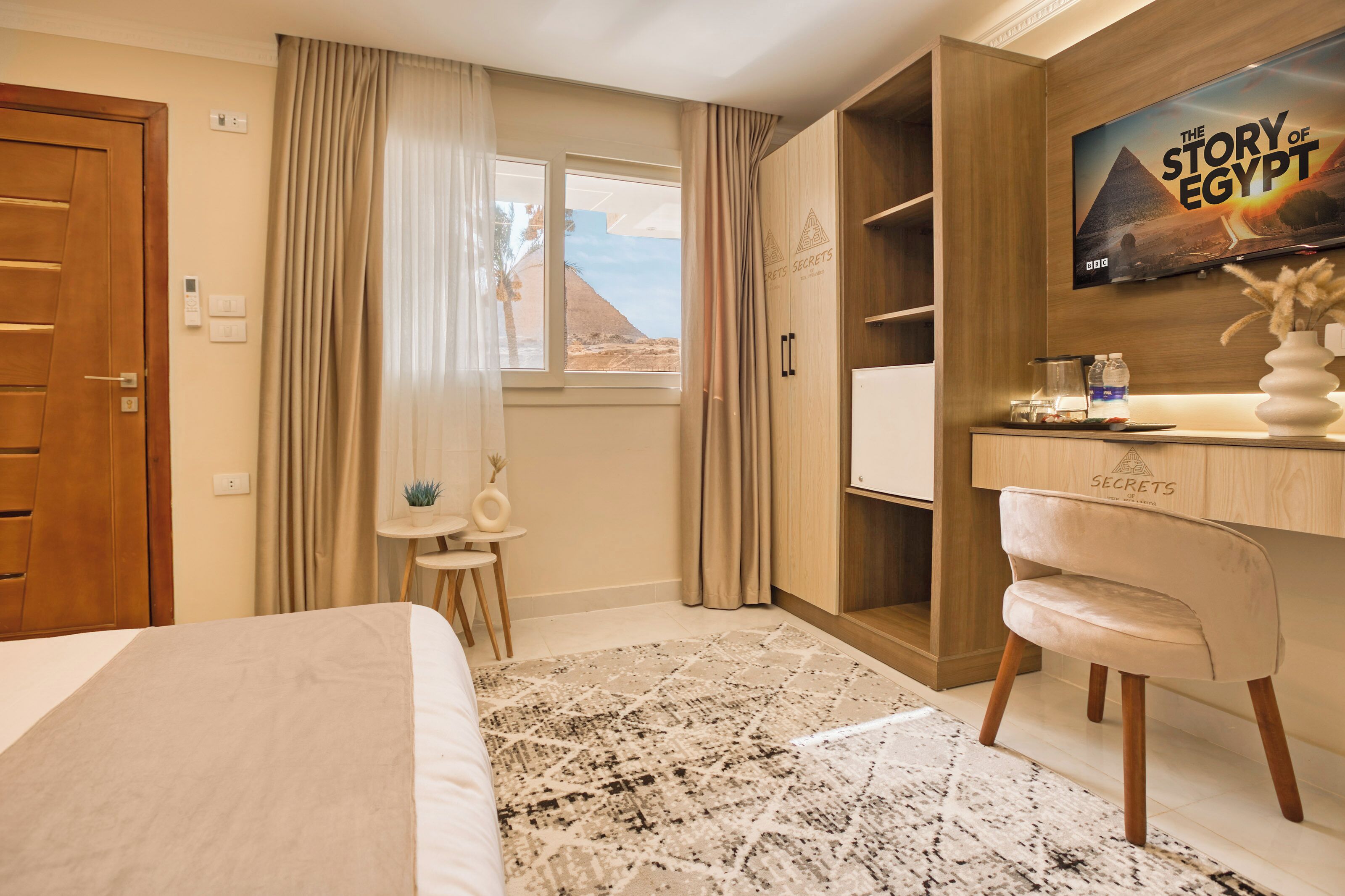 Habitación de hotel con cama, escritorio con silla, televisor montado en la pared y ventana con cortinas.