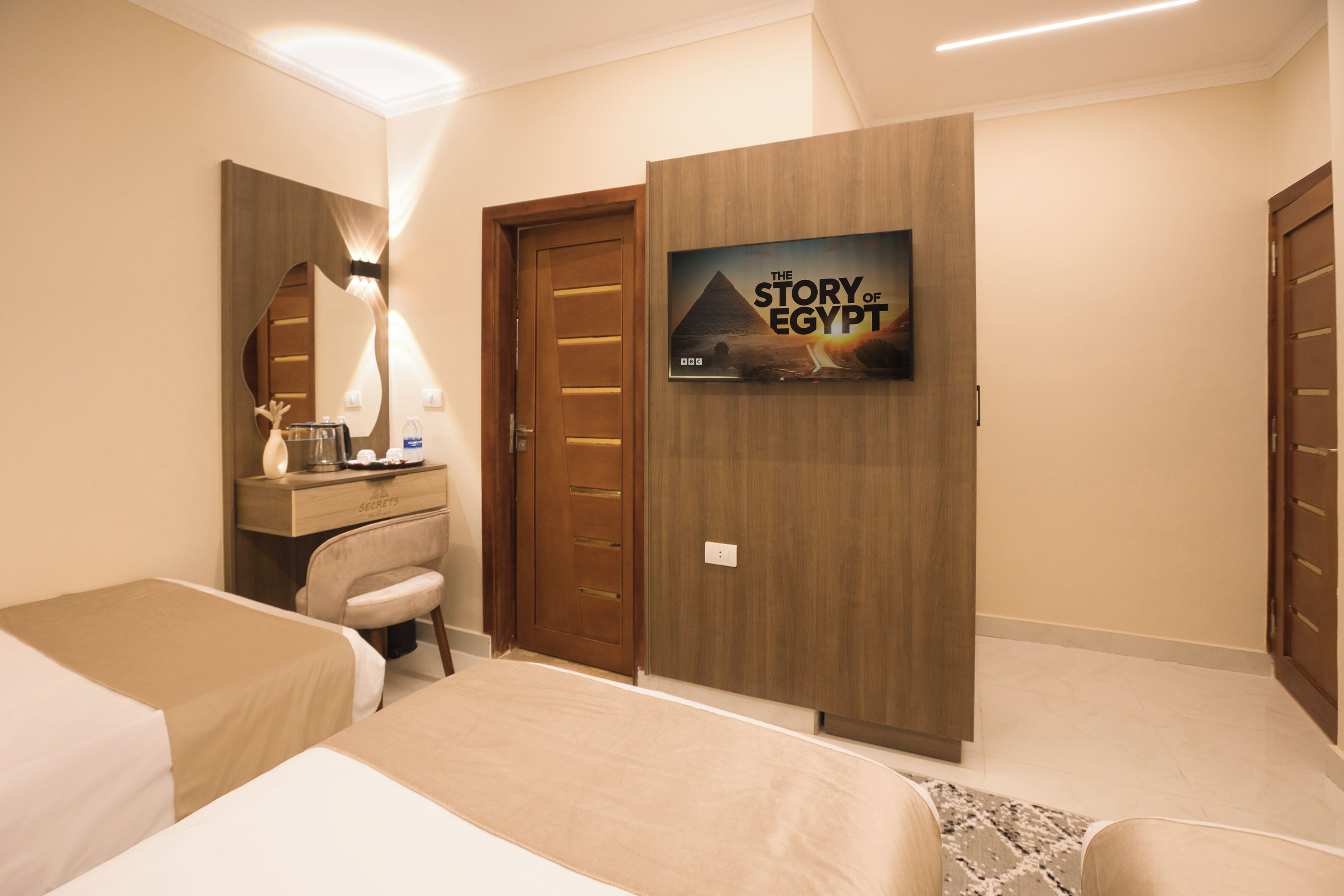 A hotel room with a bed, a desk, a chair, a wardrobe, and a wall-mounted TV displaying 'The Story of Egypt'.