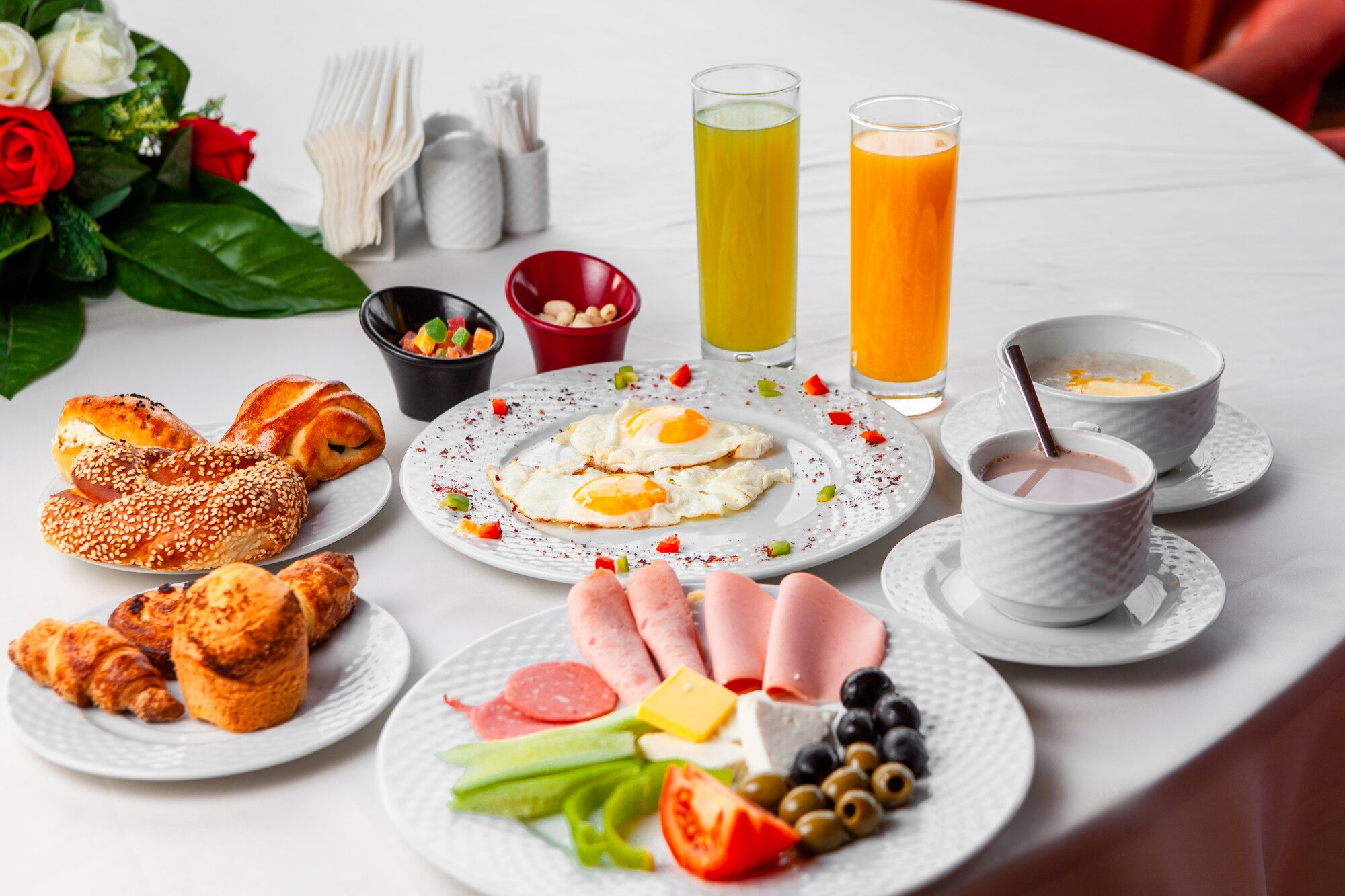 A breakfast table with croissants, eggs, ham, cheese, and various fruits. There are also two glasses of orange juice and a cup of coffee.