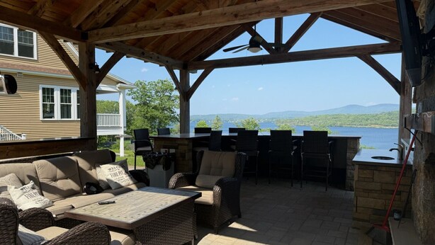 Un patio techado al aire libre con muebles de mimbre, una chimenea de piedra y vistas a un lago.