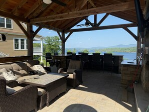 Un patio techado al aire libre con muebles de mimbre, una chimenea de piedra y vistas a un lago.