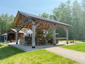 A covered outdoor seating area with stone walls and wooden beams, surrounded by a grassy lawn and trees. - Hilltop Haven on Lake Winnisquam (Sanbornton)