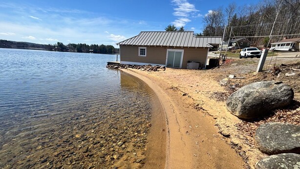 A lakeside property with a small cabin, clear water, and a sandy beach. - Lakeside A-Frame- Cozy Beach Access Retreat (Sanbornton)