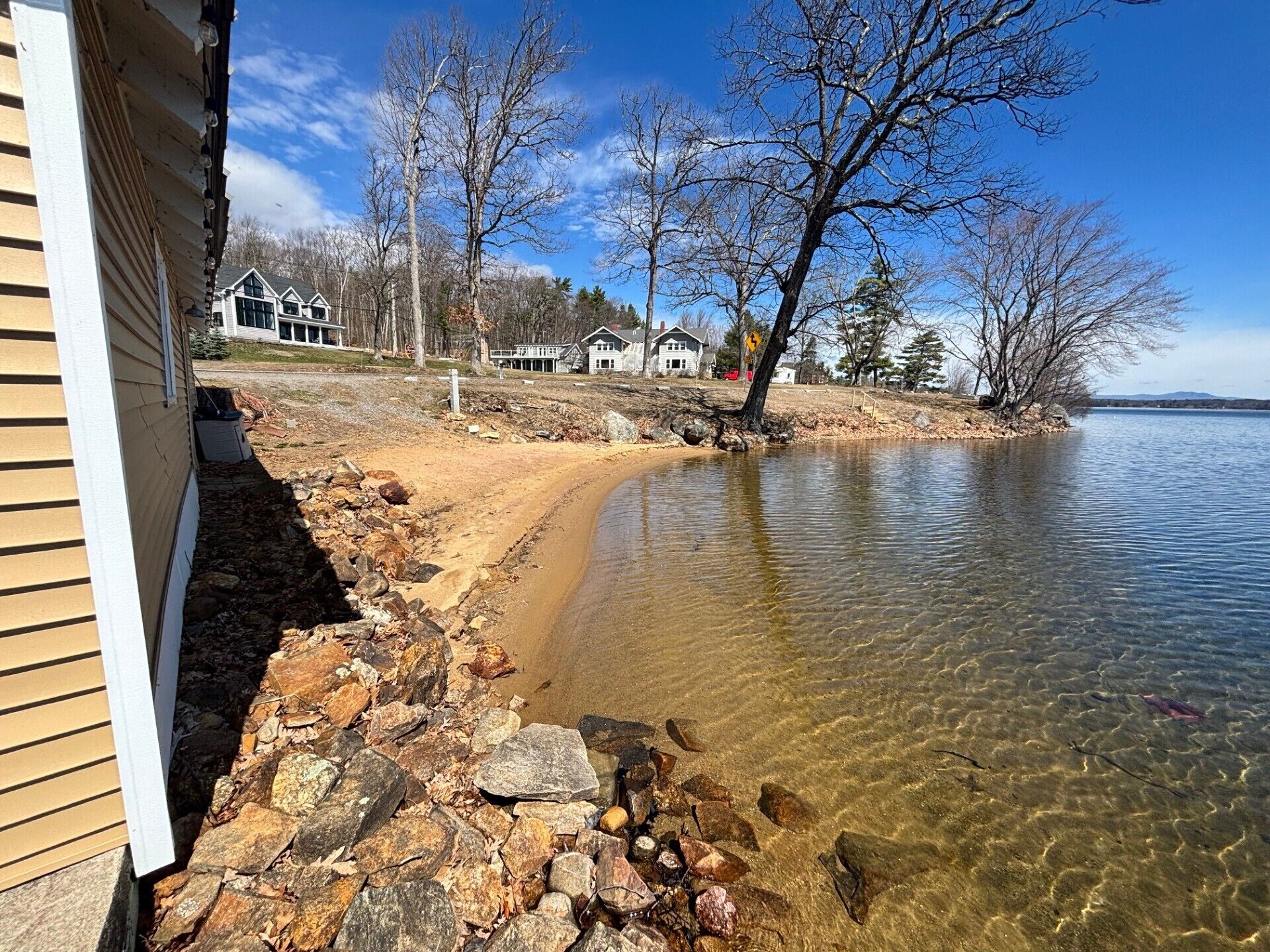A lakeside property with a clear view of the water, a sandy beach, and a row of houses in the background.