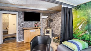 A room with a stone wall, a flat-screen TV, a fireplace, a bed with a green and blue pillow, and a large tropical mural. - Apartment 'Happy 2' with Mountain View, Private Terrace and Wi-Fi (Olsberg)