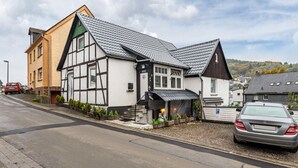 A traditional half-timbered house with a steep roof, a small porch with potted plants, and a parked car in front. - Apartment 'Happy 2' with Mountain View, Private Terrace and Wi-Fi (Olsberg)