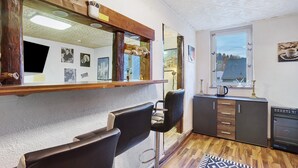 A room with a bar area, two bar stools, a wooden cabinet, a window with a view, and a patterned rug. - Apartment 'Happy' with Mountain View, Private Garden and Wi-Fi (Olsberg)
