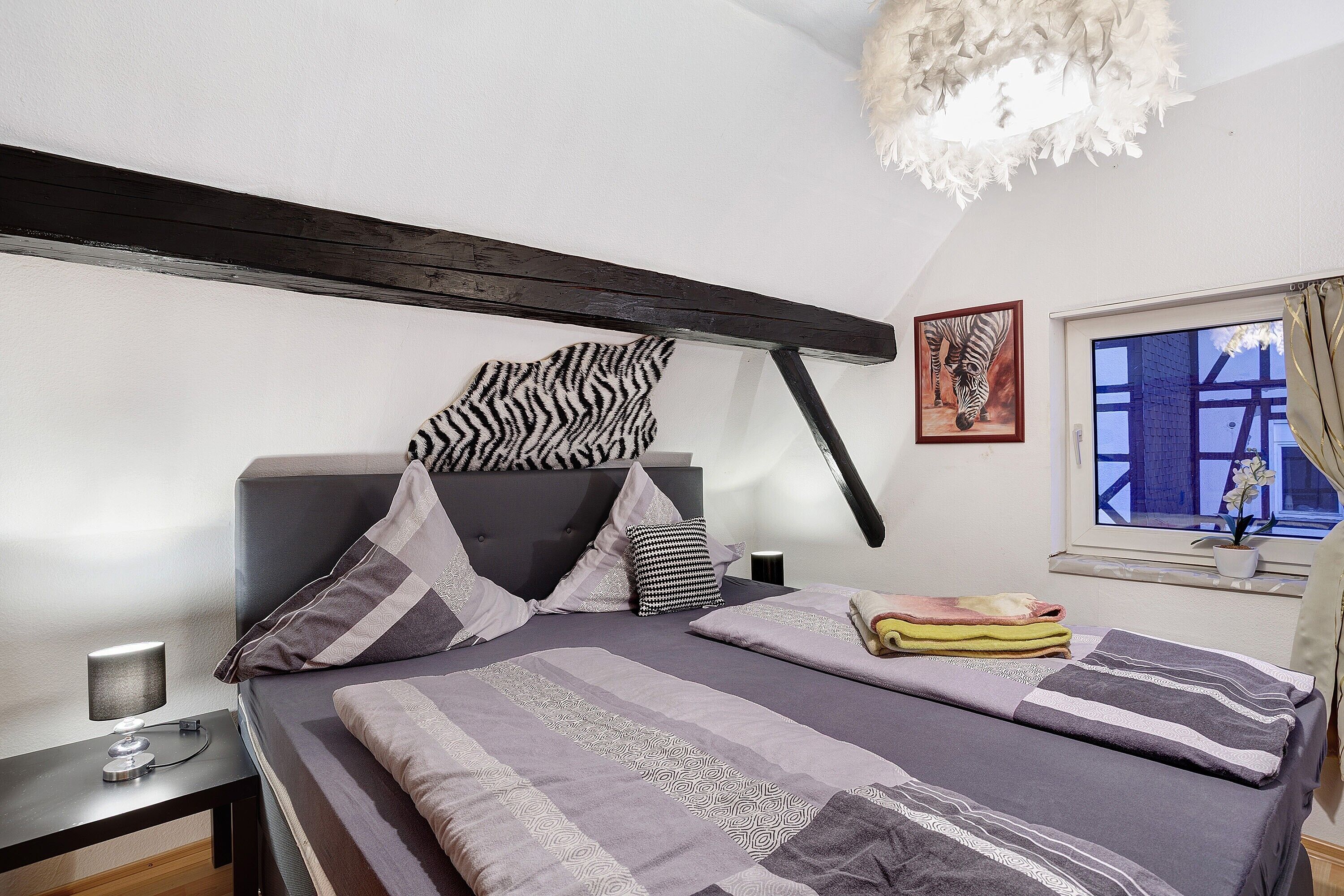 Un dormitorio con cama, almohadas, mesita de noche, lámpara, ventana con vistas y un cuadro de cebra en la pared.