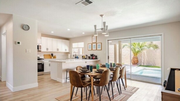 A modern kitchen with white cabinets, a dining area with a wooden table and chairs, and a large sliding glass door leading to a pool area. - Retro Modern Oasis Pool Golf (Las Vegas)
