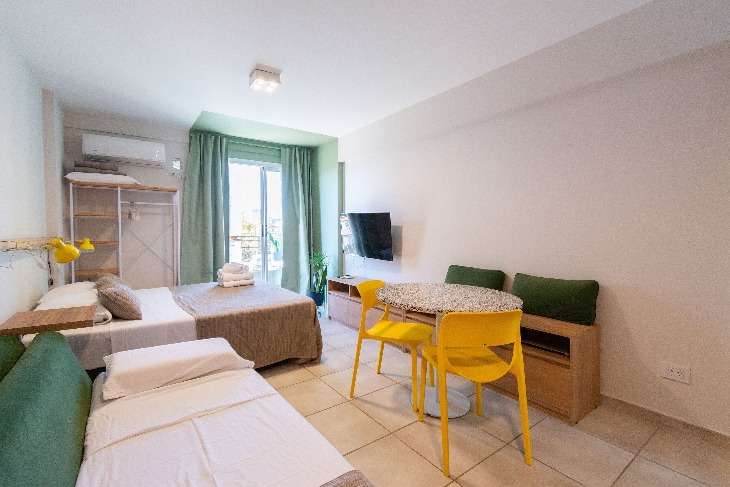 A hotel room with two beds, a dining table with yellow chairs, a TV, and a green couch.