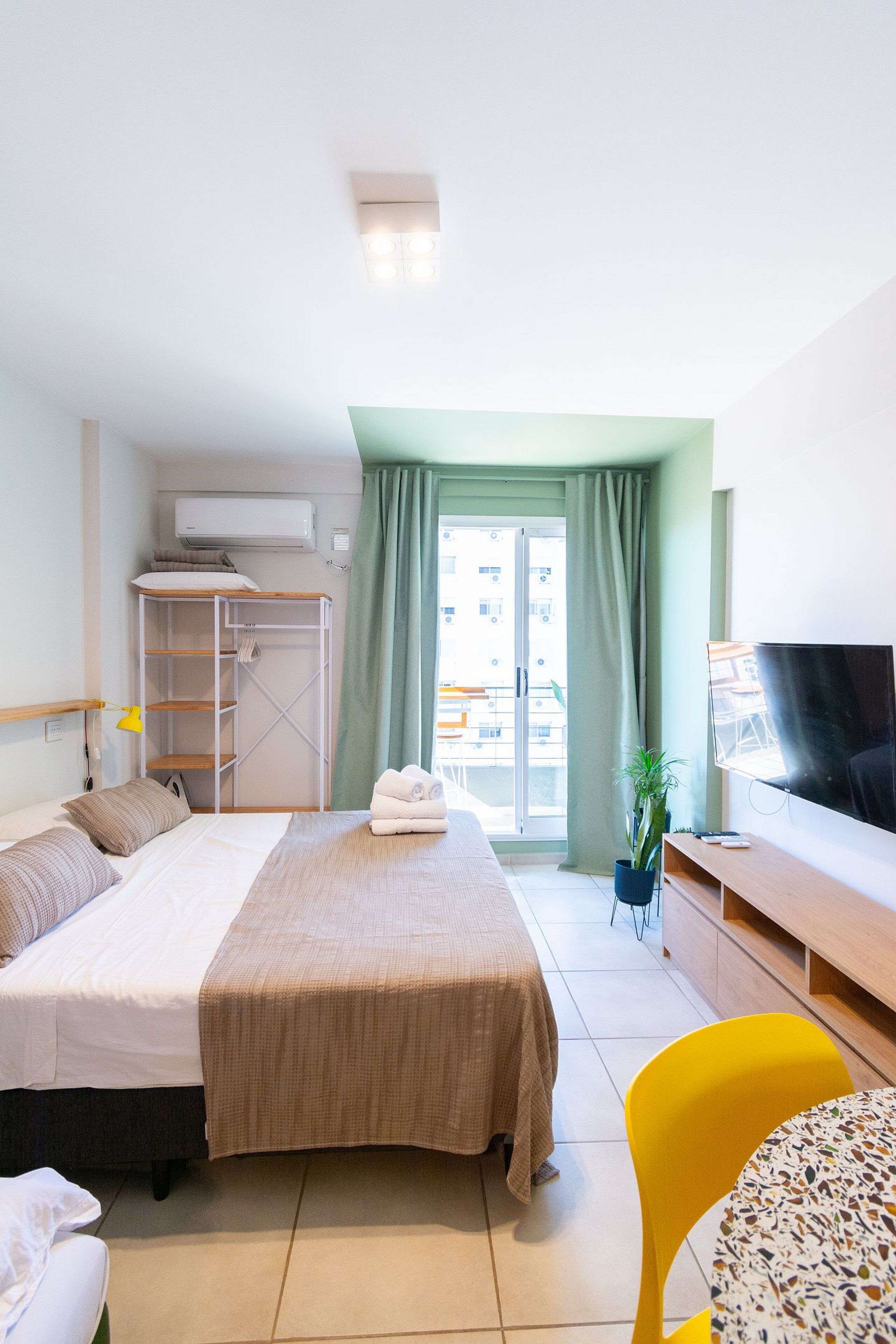 Une chambre d’hôtel avec un lit, un bureau, une télévision, une plante et un balcon donnant sur des immeubles.