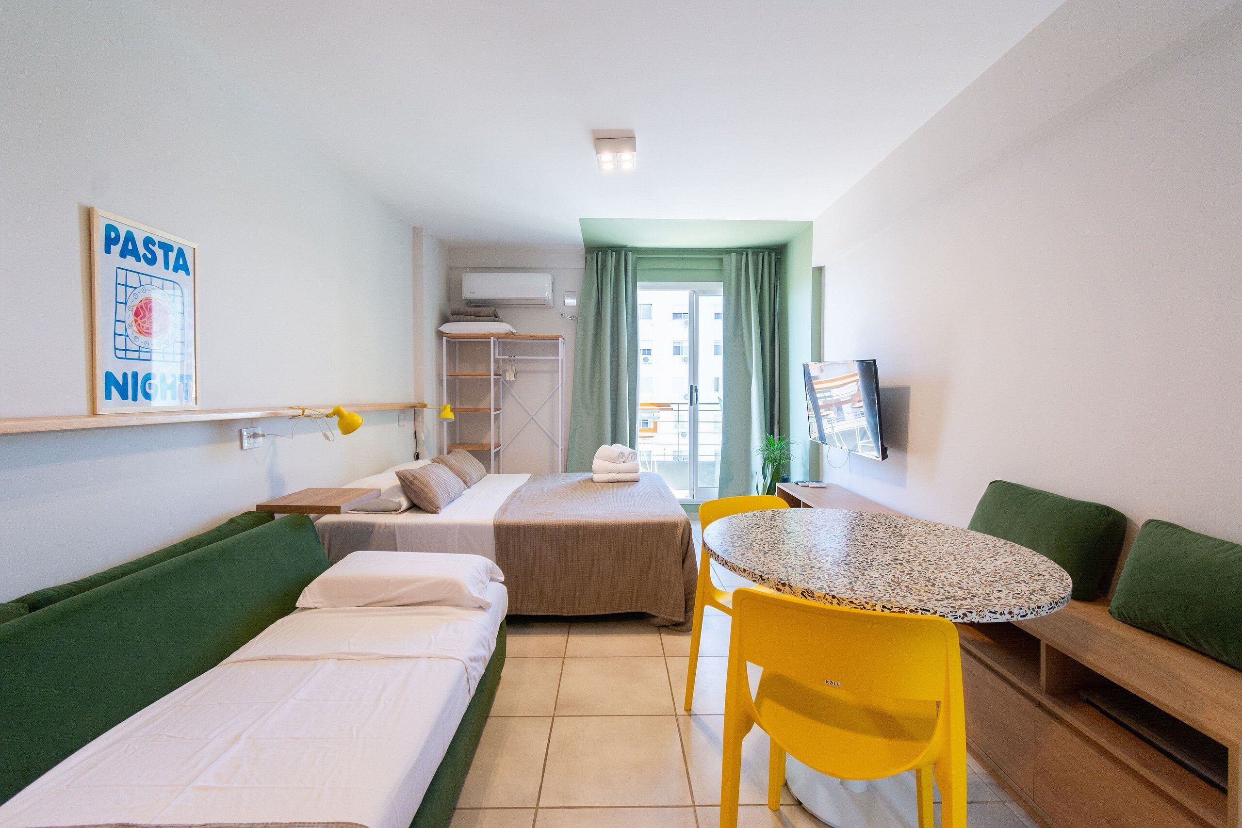 A hotel room with two single beds, a small round table with a yellow chair, a green sofa, and a TV on the wall.