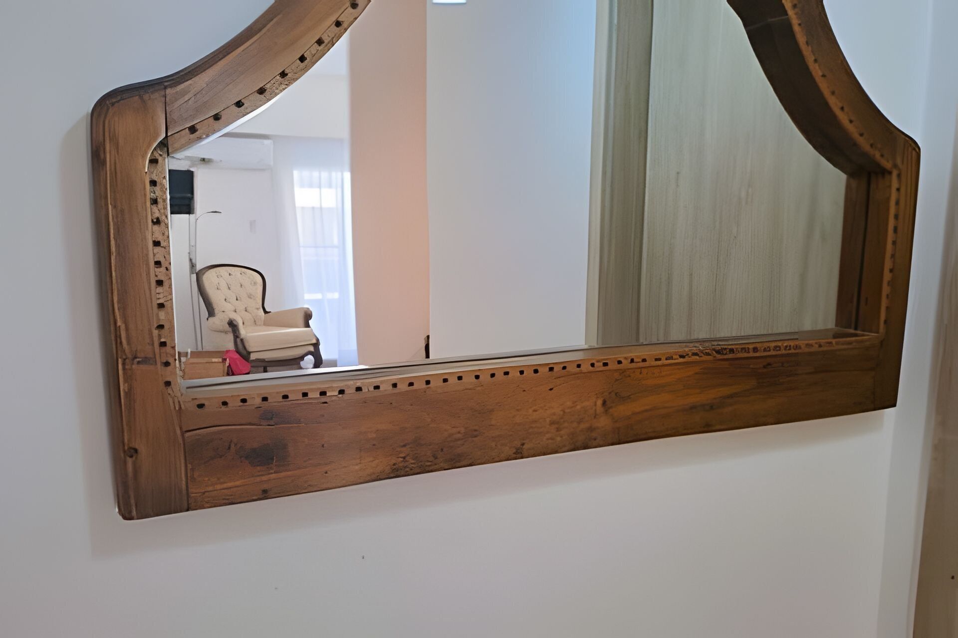 A large wooden-framed mirror with a wooden base, reflecting a room with a chair and a window.