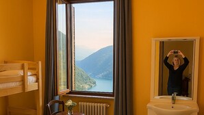 A hotel room with a yellow bed, a bunk bed, a small table with a vase, a mirror, and a view of a lake and mountains. - Baita Cao Como Lake (Brunate)