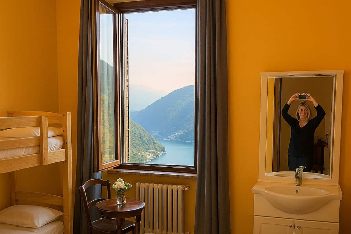 Une chambre d’hôtel avec un lit jaune, un lit superposé, une petite table avec un vase, un miroir et une vue sur un lac et des montagnes.