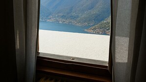 A window with sheer curtains, a view of a lake, and a distant mountain range. - Baita Cao Como Lake (Brunate)