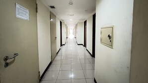 A long, narrow hallway with white tiled floors and cream-colored walls. There are doors on both sides, some with visible handles. A light fixture is mounted on the ceiling, and a security lock is visible on the right wall.