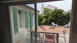 Un patio con una mesa y sillas de madera, una puerta corrediza de vidrio y vistas a un edificio y vegetación.