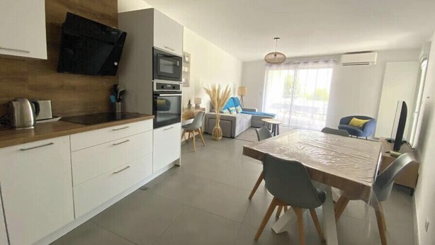 A modern kitchen with white cabinets, a wooden countertop, and a built-in oven. There is a dining area with a table and chairs, and a living space with a sofa and a TV.