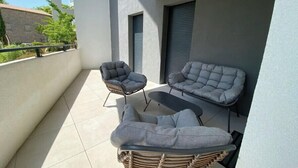 A balcony with two grey cushioned chairs and a wicker sofa, a tiled floor, and a view of a building in the background.