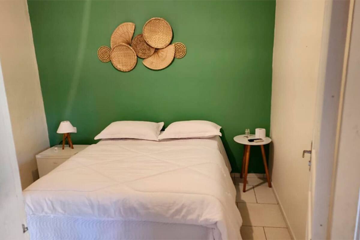 Una cama pequeña y ordenada en una habitación con paredes verdes, una mesita de noche pequeña con una lámpara, y un adorno colgado en la pared.
