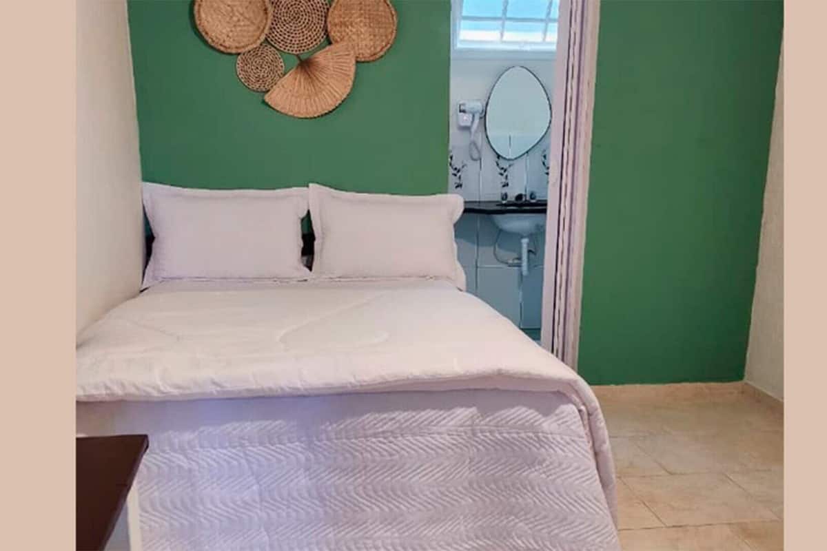 Una cama con ropa blanca, una pared verde y un baño visible a través de una puerta abierta.