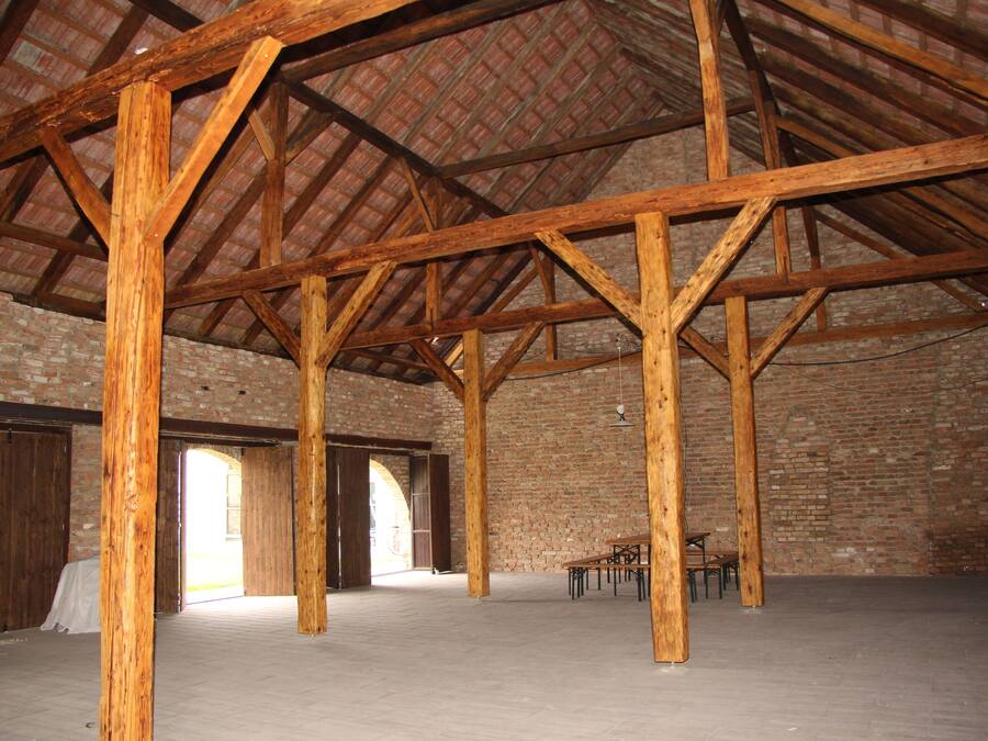 Interior con vigas de madera a la vista, paredes de ladrillo y un techo alto. Hay mesas y sillas en el centro.