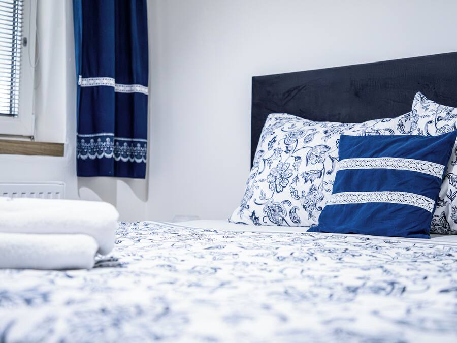 A neatly made bed with blue and white patterned bedding, a dark headboard, and a window with blinds.