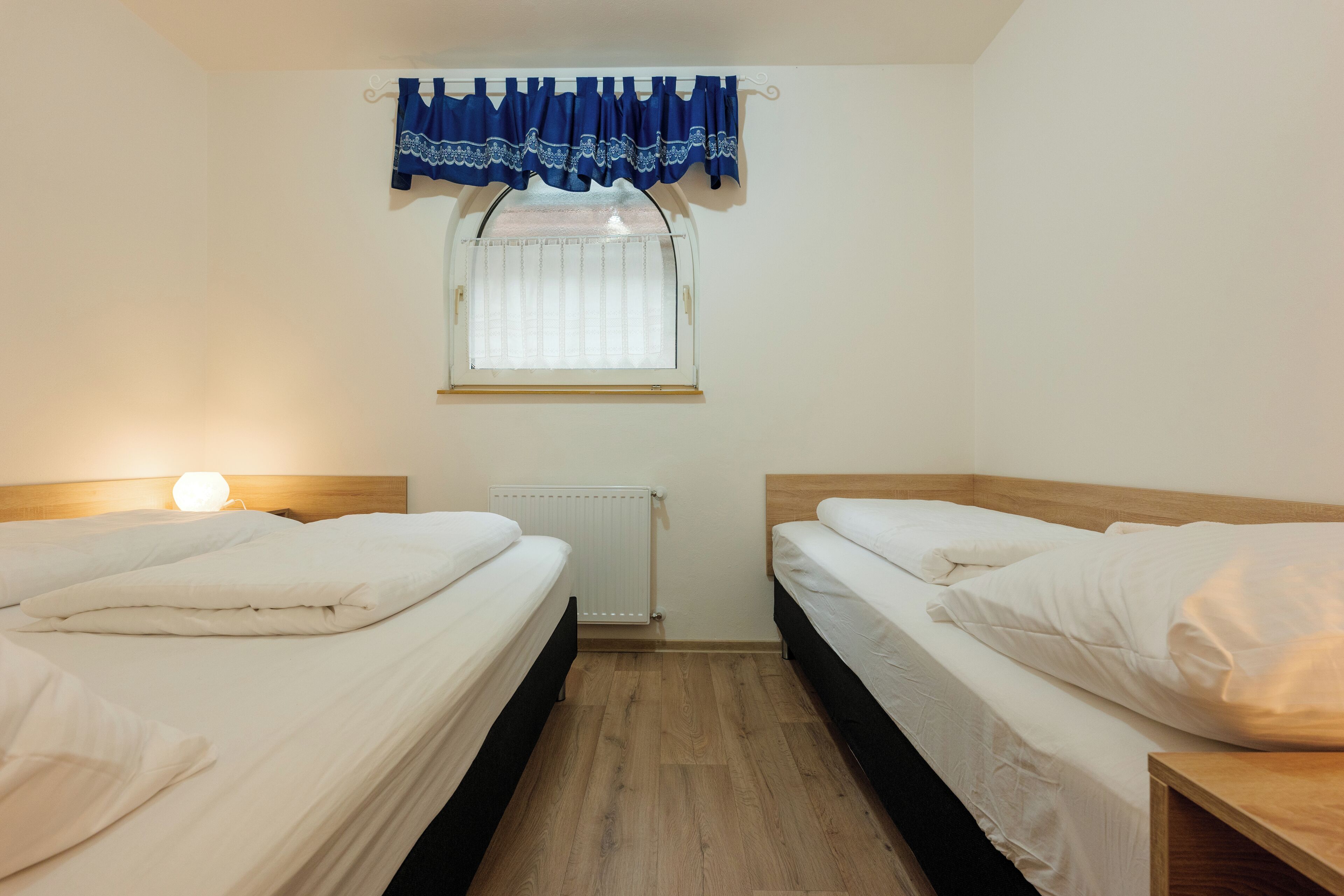 A twin room with two beds, a window with a curtain, a radiator, and a bedside table.