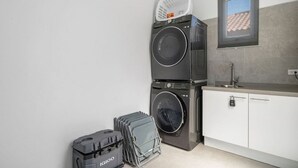 Een kleine wasruimte met gestapelde wasmachines, een klein wasbakje en een raam met uitzicht op een pannendak.
