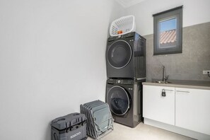 A compact laundry area with stacked washing machines, a small sink, and a window with a view of a tiled roof. - 4 BR Paradise with Private Pool! (Noord)