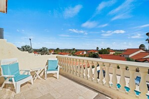 A balcony with white chairs and a table, overlooking a residential area with red-roofed houses and a clear blue sky.