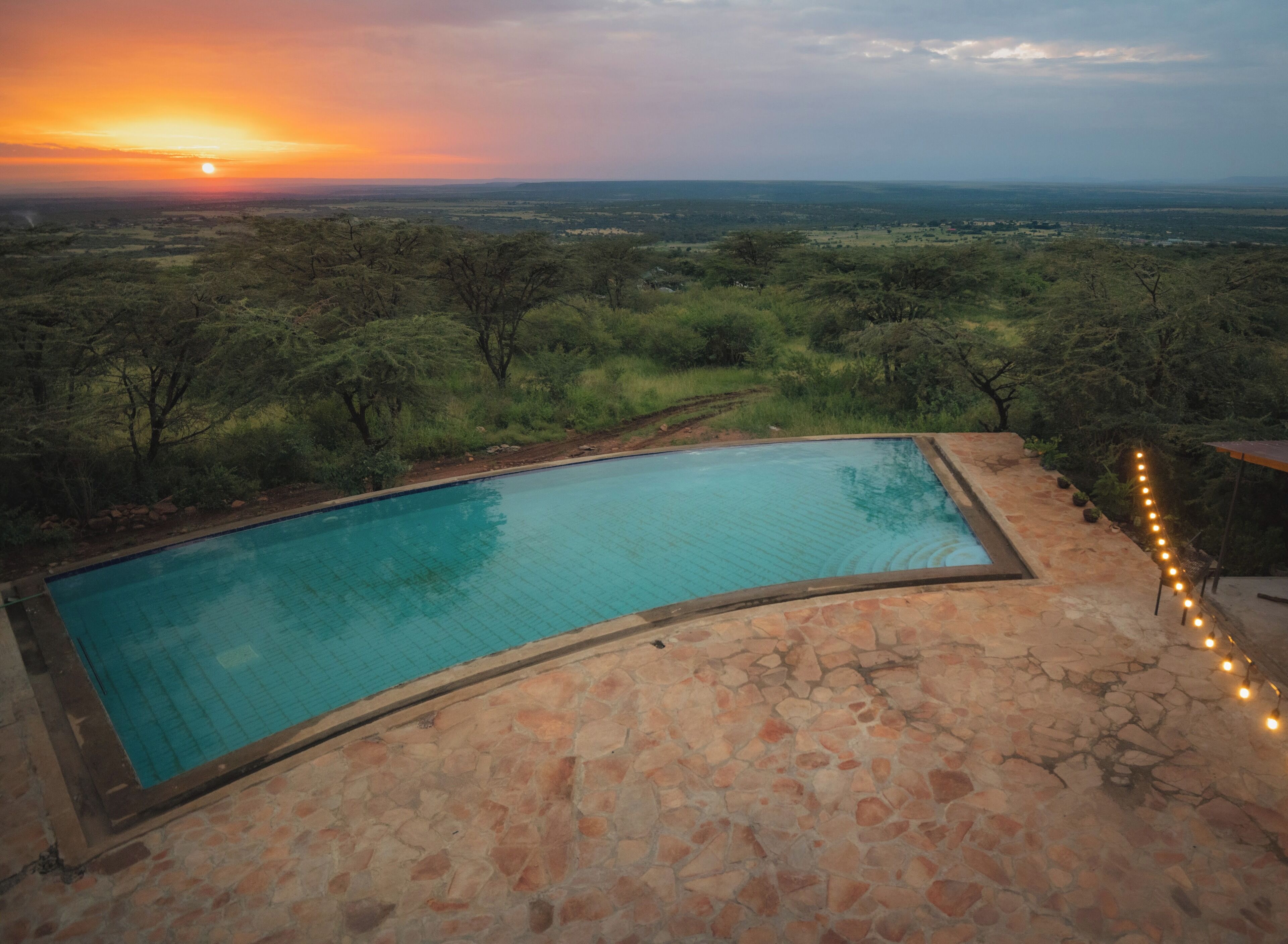 Une piscine aux bords incurvés, bordée de pierres, entourée d’un paysage arboré et offrant une vue lointaine sur l’horizon au coucher du soleil.