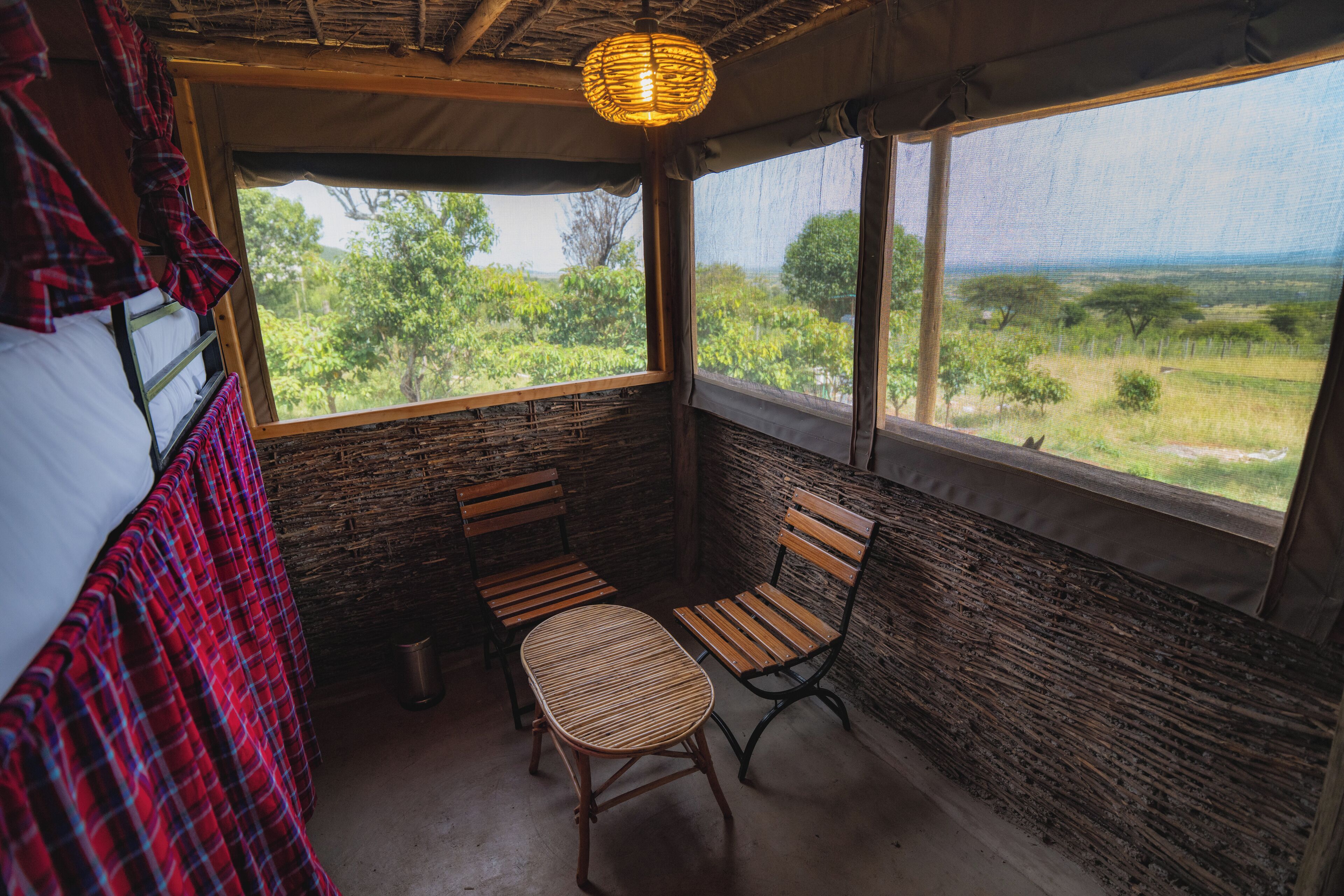 A room with a wooden table and two chairs, a window with a view of greenery, and a woven light fixture.