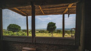 A room with a wooden bench, a thatched roof, and a view of a landscape with trees and a distant structure. - Mara hilltop (Maasai Mara)