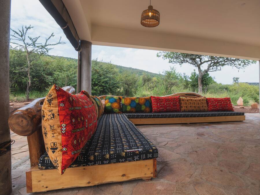 A wooden bench with colorful cushions and a patterned mat, set outdoors with a view of trees and hills.