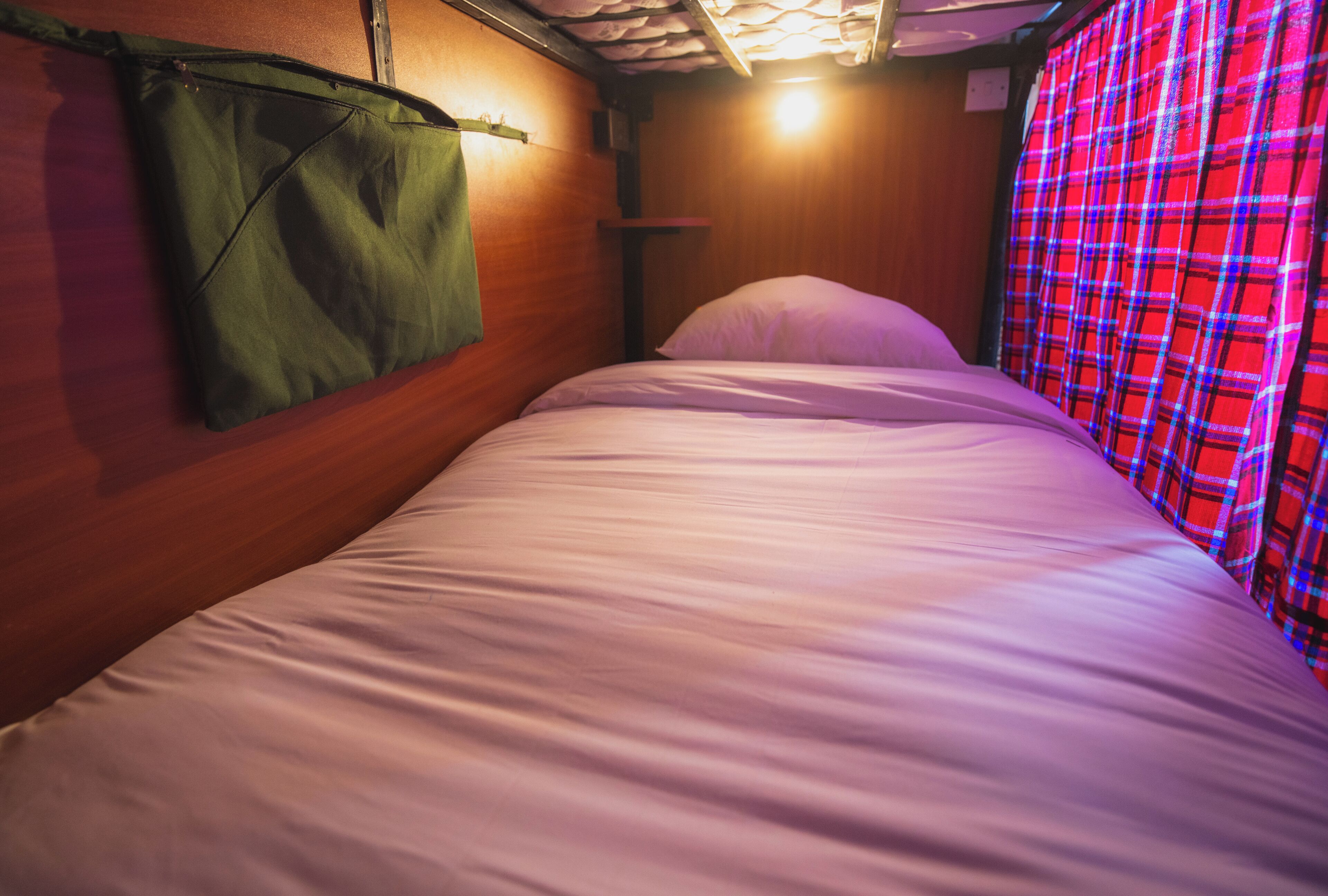 A single bed with a green bag hanging on the wall, a wooden headboard, and a plaid curtain.
