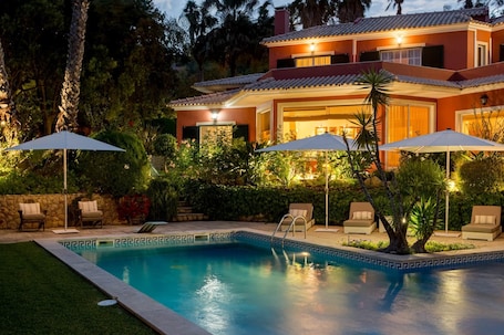 A two-story house with a swimming pool, outdoor seating, and well-lit surroundings.