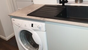 Una lavatrice compatta bianca con pannello di controllo, un lavello in acciaio inox con rubinetto nero e una finestra sopra il lavello.