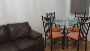 Un soggiorno con un divano in pelle marrone, un tavolo da pranzo con piano in vetro e quattro sedie, e una finestra con persiane.