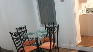 Un'area pranzo con un tavolo in vetro e quattro sedie, una finestra con persiane, un orologio appeso al muro e una porta che conduce in un'altra stanza.