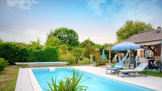 Une piscine rectangulaire dont le bord est recouvert de carreaux, entourée d’une terrasse dallée. On y trouve des chaises longues et des parasols près de la piscine, et un bâtiment avec un toit en pente en arrière-plan.