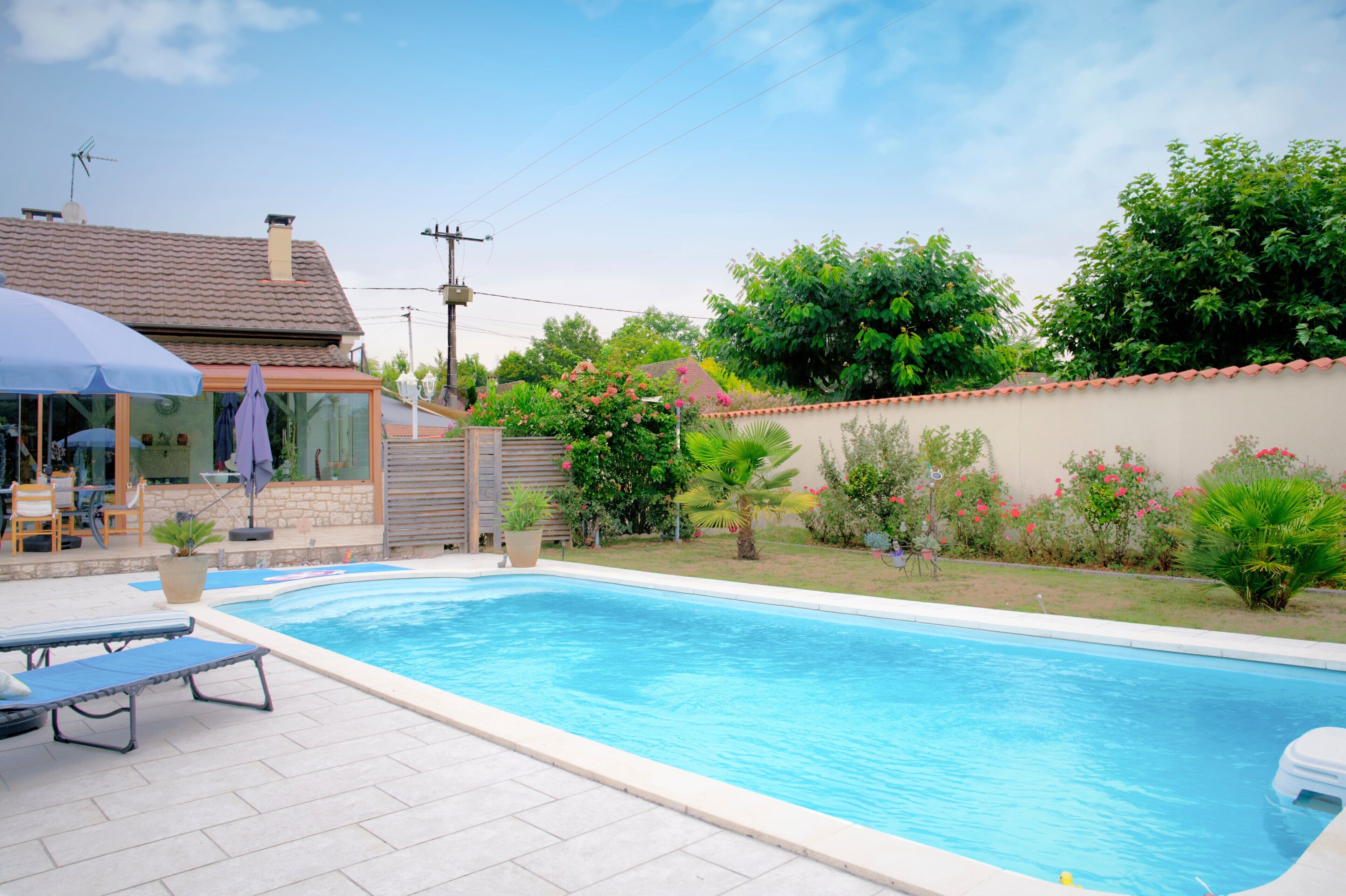 Una piscina con sillones de descanso, una zona de estar techada y un jardín con diversas plantas y flores.