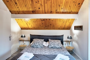 Une chambre avec un plafond en bois, un lit avec une literie à motifs, deux tables de chevet avec des lampes et un panneau décoratif fixé au mur.