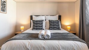 Een net opgemaakt bed met witte lakens en twee gekleurde kussens, een paar opgerolde handdoeken en twee bedlampjes op houten nachtkastjes.