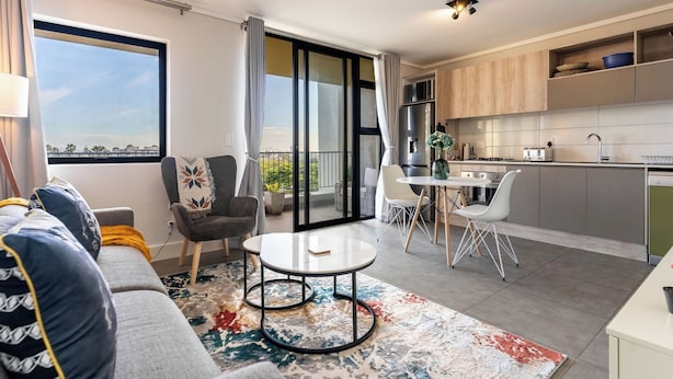 Een moderne woonkamer met een bank, een salontafel en een eethoek met stoelen. Er is een keuken met een koelkast, een gootsteen en kasten. Een groot raam biedt uitzicht op de omgeving.